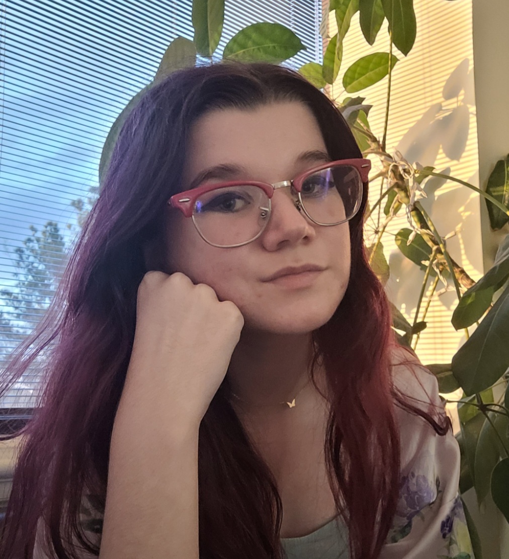 A white woman with long brown hair and red cateye glasses, resting her chin on her fist and smiling at the camera.